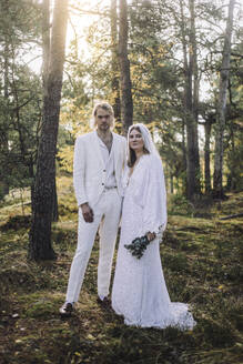Porträt von gut gekleideten Braut und Bräutigam stehen durch Bäume im Wald bei der Hochzeit - MASF35750