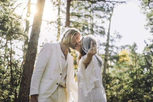 Bride showing ring finger while kissing groom on mouth in forest during wedding - MASF35753