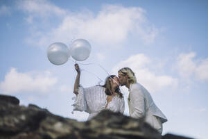Romantische Braut hält weiße Heliumballons und küsst den Bräutigam gegen den Himmel - MASF35763