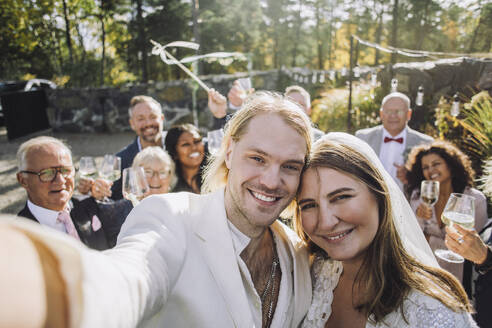 Ein frisch verheiratetes Paar fängt Erinnerungen an seinen freudigen Hochzeitstag mit Gästen im Hintergrund ein. - MASF35766