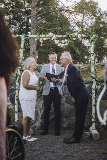 Älteres Paar tauscht das Eheversprechen aus, während der Hochzeitszeremonie steht der Pfarrer an einer Wand mit Blumenschmuck - MASF35780