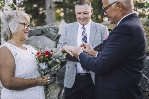 Senior groom putting wedding ring in bride's finger by minister during ceremony - MASF35781