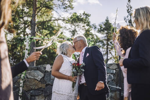 Ältere Braut mit Blumenstrauß küsst Bräutigam auf den Mund inmitten der Gäste während der Hochzeitsfeier - MASF35782