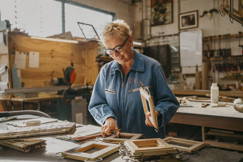 Smiling senior carpenter examining frames at workshop - MASF35813