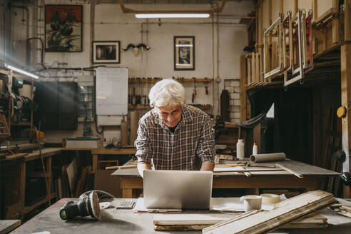 Älterer männlicher Tischler bei der Arbeit an einem Laptop in einer Reparaturwerkstatt - MASF35832