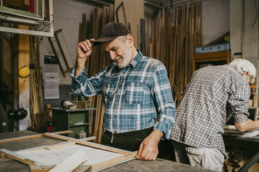 Senior male carpenter adjusting cap while working with colleague at repair shop - MASF35838