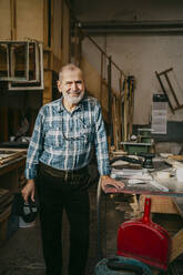Lächelnder männlicher Zimmermann, der in der Nähe einer Werkbank in einer Reparaturwerkstatt steht - MASF35854