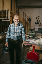 Smiling male carpenter standing near workbench at repair shop - MASF35854