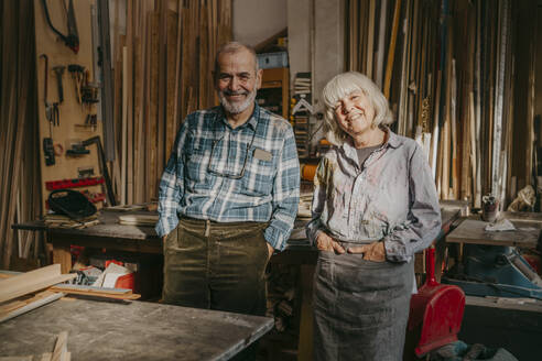Portrait of happy male and female carpenters with hands in pockets standing at workshop - MASF35859