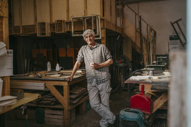 Smiling senior craftsman leaning on workbench at carpentry workshop - MASF35861