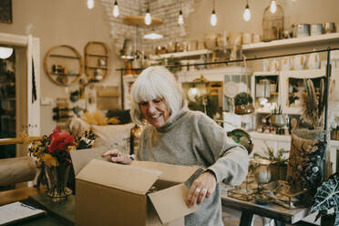Happy senior female entrepreneur opening cardboard box at interior shop - MASF35873