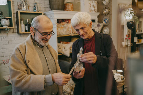 Senior male customer checking price tag of glass figurine held by owner at store - MASF35891