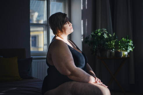 Depressed lonely fat woman sitting and looking through the window at home at dark. - HPIF07067