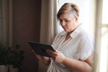 An overweight woman in white shirt standing by window and using tablet. - HPIF07073