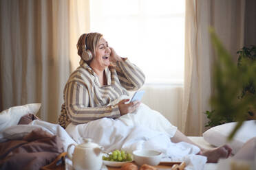 Eine glückliche übergewichtige Frau mit Kopfhörern und Smartphone frühstückt zu Hause im Bett. - HPIF07082