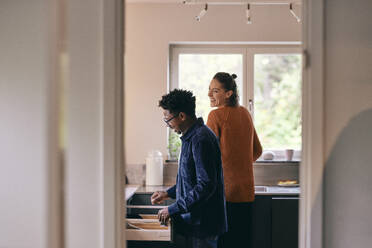 Happy multiracial couple helping each other while standing in kitchen at home - MASF35995