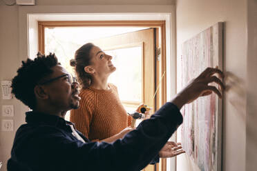 Paar, das sich gegenseitig beim Aufhängen eines Gemäldes an der Wand in der Nähe der Tür zu Hause unterstützt - MASF36000
