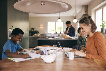 Happy mother using laptop with son coloring in book while sitting at dining table in home - MASF36001