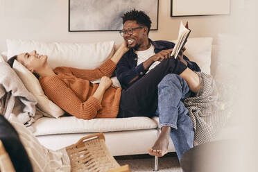 Happy multiracial couple having fun while reading books on sofa at home - MASF36012
