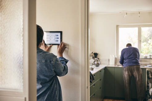Rückansicht einer Frau, die ein digitales Tablet benutzt, das zu Hause an der Wand befestigt ist - MASF36047