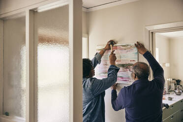 Rückansicht eines älteren Mannes, der einer Frau beim Aufhängen eines Gemäldes an der Hauswand hilft - MASF36051