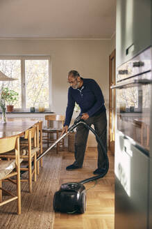 Älterer Mann reinigt Teppich mit Staubsauger im Stehen zu Hause - MASF36056