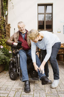 Ältere Frau in voller Länge, die einem Mann mit Behinderung hilft, der in einem motorisierten Rollstuhl im Hinterhof sitzt - MASF36073