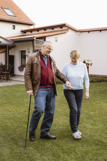 Senior man holding hand of woman while walking with cane at lawn in back yard - MASF36074