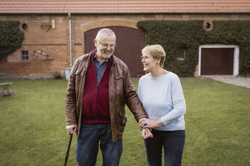 Lächelnde ältere Frau hält die Hand eines Mannes, der mit einem Stock auf einer Wiese im Hinterhof geht - MASF36077