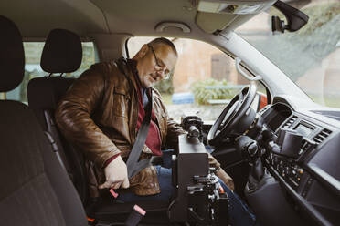 Senior man with disability fastening seat belt while sitting inside van - MASF36081