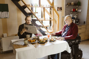 Young man showing newspaper to retired senior father with disability while eating breakfast at home - MASF36090