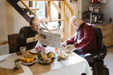 Junger Mann zeigt seinem älteren Vater mit Behinderung beim Frühstück zu Hause die Zeitung - MASF36091