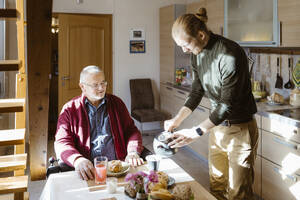 Junger männlicher Hausmeister gießt Kaffee für einen älteren Mann im Ruhestand mit Behinderung beim Frühstück zu Hause ein - MASF36092