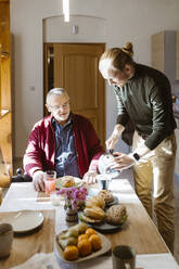 Young man pouring coffee for retired senior father with disability during breakfast at home - MASF36093