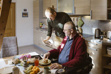 Young caretaker sharing newspaper with senior man while eating breakfast at home - MASF36095