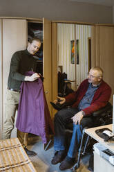 Junge männliche Pflegekraft zeigt einem älteren Mann mit Behinderung, der zu Hause im motorisierten Rollstuhl sitzt, ein Hemd - MASF36098