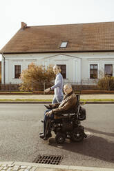 Seitenansicht eines älteren Mannes mit Behinderung in einem motorisierten Rollstuhl, der von einer jungen Betreuerin auf einer Straße an einem sonnigen Tag begleitet wird - MASF36099