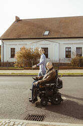 Side view of senior man with disability in motorized wheelchair by young caregiver walking on road during sunny day - MASF36099