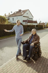 Lächelnder älterer Mann mit Behinderung in einem motorisierten Rollstuhl, der von einer jungen Betreuerin bei Sonnenschein auf der Straße geführt wird, in voller Länge - MASF36101