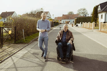 Älterer Mann mit Behinderung in vollem Umfang im motorisierten Rollstuhl mit junger Betreuerin auf der Straße bei sonnigem Wetter - MASF36102