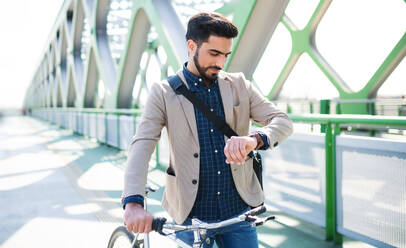 Portrait of young business man commuter with bicycle going to work outdoors in city, checking the time. - HPIF07088