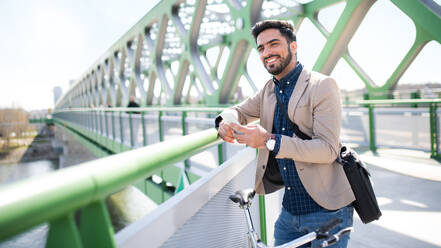 Porträt eines jungen Geschäftsmannes, der mit dem Fahrrad zur Arbeit in der Stadt fährt und auf einer Brücke sein Smartphone benutzt. - HPIF07090