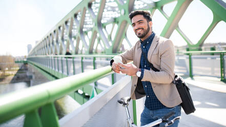 Portrait of young business man commuter with bicycle going to work outdoors in city, using smartphone on bridge. - HPIF07090