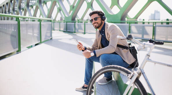 Porträt eines jungen Geschäftsmannes, der mit dem Fahrrad zur Arbeit in der Stadt fährt und dabei sein Smartphone benutzt. - HPIF07094