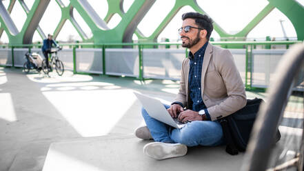 Portrait of young business man commuter with bicycle on the way to work outdoors in city, using laptop. - HPIF07096