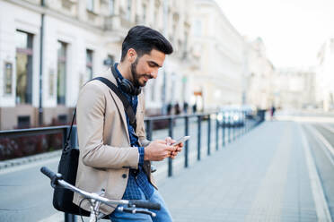 Porträt eines jungen Geschäftsmannes, der mit dem Fahrrad zur Arbeit in der Stadt fährt und dabei sein Smartphone benutzt. - HPIF07100