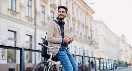 Porträt eines jungen Geschäftsmannes, der mit dem Fahrrad zur Arbeit in der Stadt fährt und dabei sein Smartphone benutzt. - HPIF07101