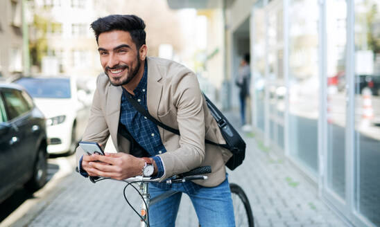 Porträt eines jungen Geschäftsmannes, der mit dem Fahrrad zur Arbeit in der Stadt fährt und dabei sein Smartphone benutzt. - HPIF07103