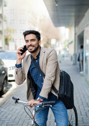 Porträt eines jungen Geschäftsmannes, der mit dem Fahrrad und seinem Smartphone zur Arbeit in der Stadt fährt. - HPIF07104