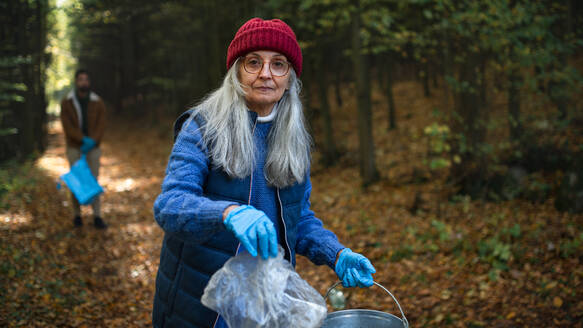 A senior woman volunteer looking at camera and cleaning up forest from waste, community service concept - HPIF07111